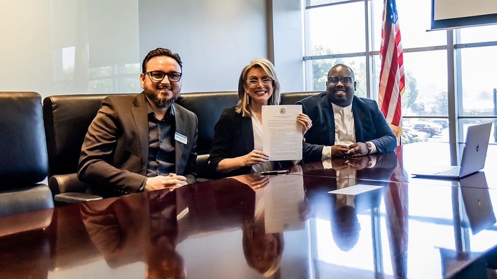 dr. ruarte, president angie, + jason fletcher smiling + holding up the signing papers confirming partnership