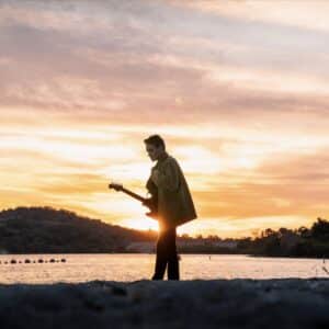 a photo of calvin holding. a guitar near a lake with the sunset in the background