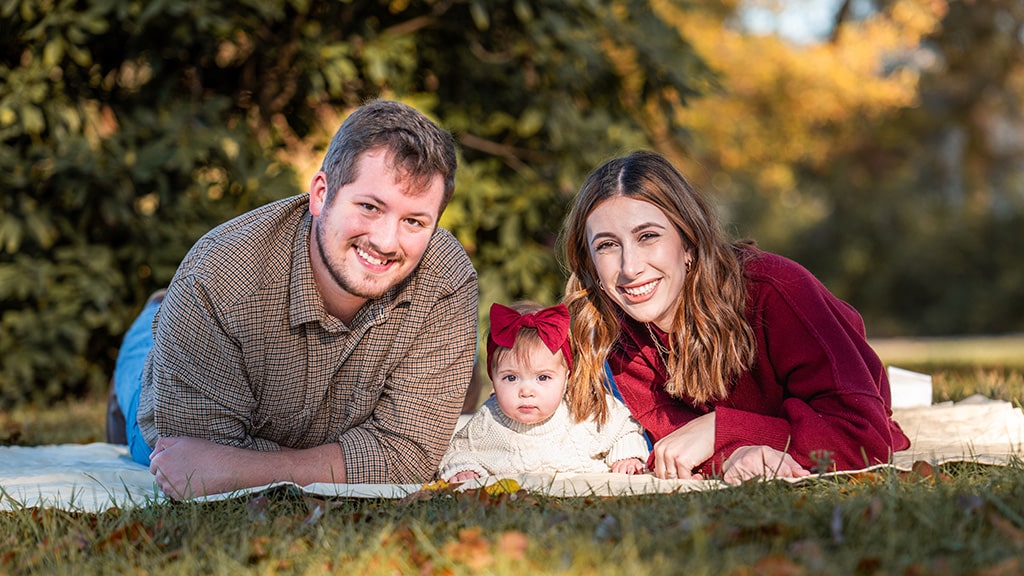 a photo of shea, her husband, and daughter smiling and laying down on the grass in a group photo