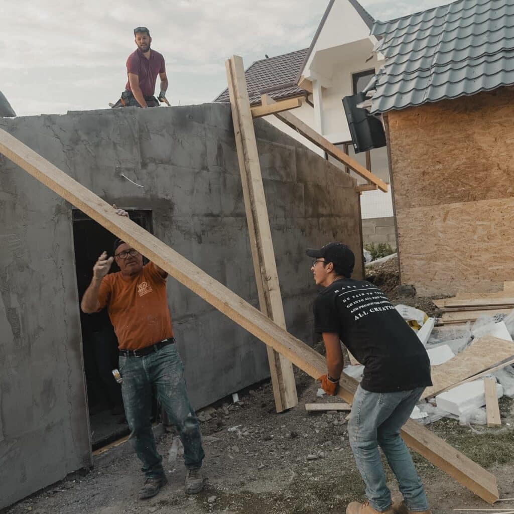 a male student lifting a plank of wood onto the roof of the house being built